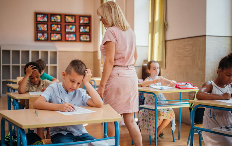 Fotografía de un aula con alumnos y una maestra de pie entre ellos.